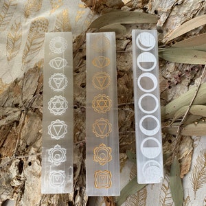 May include: Three white selenite sticks with gold etched designs. The left stick has seven chakra symbols, the middle stick has seven chakra symbols, and the right stick has five moon phases.