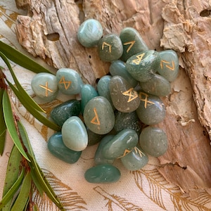 Puede incluir: Un conjunto de piedras de runas de aventurina verde con runas doradas. Las piedras son pulidas y lisas, y est&aacute;n dispuestas sobre un trozo de corteza.