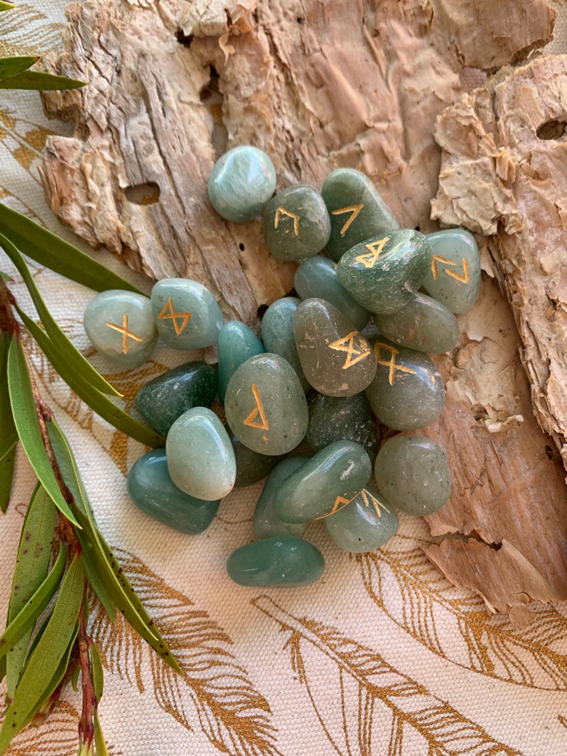 Puede incluir: Un conjunto de piedras de runas de aventurina verde con runas doradas. Las piedras son pulidas y lisas, y est&aacute;n dispuestas sobre un trozo de corteza.