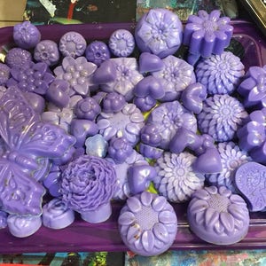Puede incluir: Una colección de jabones de color morado y lavanda en varias formas, incluyendo flores, mariposas y diseños geométricos. Los jabones están dispuestos en una bandeja morada.