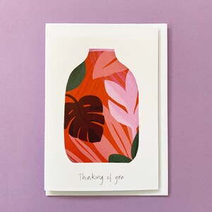 May include: A white card featuring an illustration of a vase with a red, pink and green floral pattern. The text 'Thinking of you' is written in black ink at the bottom of the card.