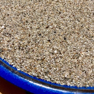 May include: Close-up of a blue ceramic bowl filled with fine, light-colored sand. The sand is composed of tiny, irregularly shaped grains in shades of beige, gray, and white. The bowl's rim is a vibrant blue.