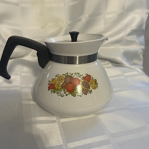 Op de afbeelding: Witte koffiepot met een zwarte handgreep en deksel. De pot heeft een zilveren band in het midden en een bloemenontwerp met gele, oranje en bruine elementen. De pot is waarschijnlijk gemaakt van keramiek of email.