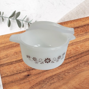 May include: White glass casserole dish with a lid, featuring a brown floral pattern around the rim. The dish has two handles and is set on a wooden surface. A sprig of greenery is in the background.
