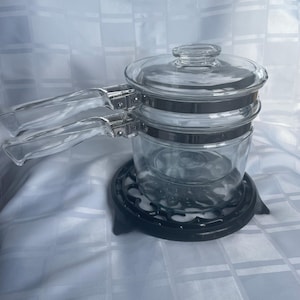 Può includere: Una pentola di vetro trasparente con coperchio e due manici. La pentola poggia su un sottopentola in metallo nero. La pentola è realizzata in vetro trasparente e ha una fascia metallica argentata al centro. Anche i manici sono in vetro trasparente.