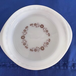 Może przedstawiać: Białe, okrągłe naczynie Pyrex z brązowym kwiatowym wzorem. Naczynie ma falisty brzeg i dwa małe uchwyty. Wzór kwiatowy jest ułożony w okrąg pośrodku naczynia.