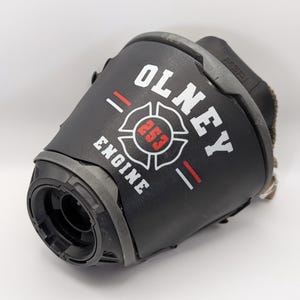 Könnte beinhalten: Ein schwarzes Feuerwehrhelm-Frontschild mit dem weißen Schriftzug OLNEY und ENGINE sowie einem zentralen roten Malteserkreuz-Emblem mit der Nummer 253. Das Design enthält zwei horizontale rote Linien neben dem zentralen Emblem.