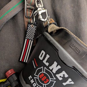 May include: A black firefighter mask bag with the text OLNEY ENGINE and the number 253 inside a Maltese cross emblem. A rectangular tag featuring a black and white American flag with a single horizontal red stripe hangs from a metal clip.