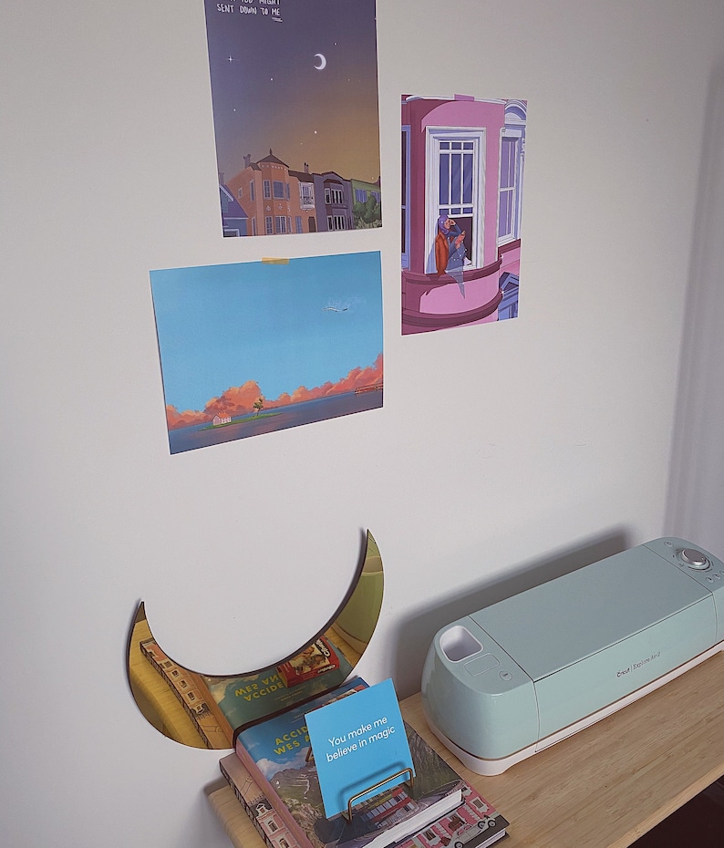 Pu&ograve; includere: Una macchina da taglio artigianale di colore azzurro chiaro con un bordo dorato su una superficie di legno. Un decoro da parete a forma di mezzaluna dorata si trova sul lato sinistro dell'immagine. Una pila di libri con un biglietto blu che dice "You make me believe in magic" si trova davanti alla macchina.