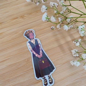 May include: A sticker of a cartoon woman wearing a black dress, a gold sparkly top, a black hat, and black shoes with gold accents. She is holding a brown briefcase. The sticker is on a wooden background.