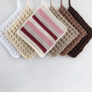 Può includere: Una collezione di sei presine all'uncinetto fatte a mano in vari colori. Le presine sono di forma quadrata con un punto waffle strutturato. I colori includono bianco, beige, rosa, bordeaux, marrone e marrone scuro. Ogni presina ha un anello per appenderla.