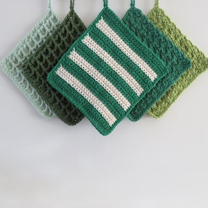 Puede incluir: Cinco agarradores de cocina cuadrados de ganchillo en varios tonos de verde. El agarrador central presenta rayas horizontales blancas sobre un fondo verde oscuro, mientras que los otros muestran patrones texturizados en verde claro, oliva y bosque.