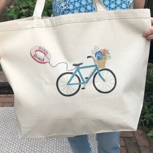 May include: A white canvas tote bag with a blue bicycle with a basket of flowers and a pink life preserver with the text "Beach Bound".