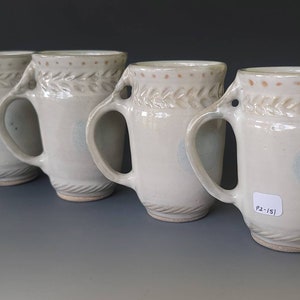 May include: Four white ceramic mugs with a light brown glaze and a leaf design around the top. The mugs have two handles and a light blue glaze on the bottom.