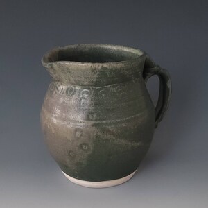 May include: A green ceramic pitcher with a white rim and a handle. The pitcher has a mottled glaze and a decorative design around the top.