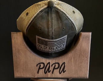 Puede incluir: Un soporte para sombreros de madera con una base de metal negro. El soporte está grabado con la palabra "PAPA" y presenta una gorra de béisbol verde y beige con un parche de "Old Glory Rustic Sign Co.".