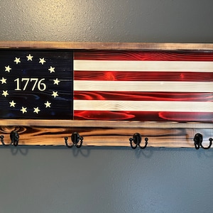 May include: A wooden wall-mounted coat rack with a distressed finish. The rack features a framed American flag with 13 stars and the year 1776. The rack has four hooks for hanging items.