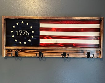 May include: A wooden wall-mounted coat rack with a distressed finish. The rack features a framed American flag with 13 stars and the year 1776. The rack has four hooks for hanging items.