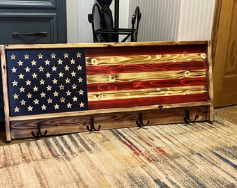 May include: A wooden coat rack with a distressed finish features a burnt wood American flag design. The rack has three hooks and is perfect for hanging coats, hats, or bags.
