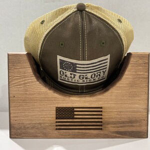 May include: A wooden hat rack with a brown finish. The rack has a curved top to hold a baseball cap. The cap is green with a tan mesh back and a white patch that says "Old Glory Rustic Sign Co.". The rack has an American flag design engraved on the front.
