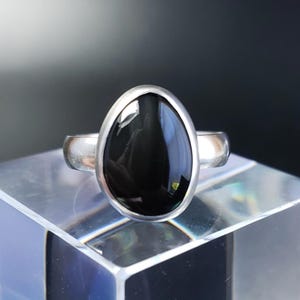 May include: A silver ring featuring a large, oval-shaped black stone. The stone is set in a silver bezel, and the band is a simple, polished silver. The ring is displayed on a clear, geometric stand against a dark background.