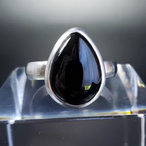 May include: A silver ring with a teardrop-shaped black stone. The stone is set in a silver bezel and has a polished, reflective surface. The ring is displayed on a clear, glass-like surface.