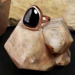 May include: A black gemstone ring with a copper setting. The ring is sitting on a light brown rock.