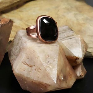 Puede incluir: Un anillo de cobre con una piedra preciosa negra engastada en un bisel cuadrado. El anillo está sobre un cristal blanco.