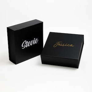 May include: Two black gift boxes with lids. The box on the left has the name "Stevie" printed in white lettering. The box on the right has the name "Jessica" printed in gold lettering.