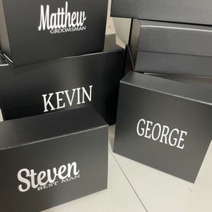 May include: Four black gift boxes with white text. The boxes are stacked on top of each other. The text on the boxes reads "Matthew Groomsman", "Kevin", "George", and "Steven Best Man".