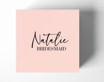May include: A light pink card with the name "Natalie" written in black cursive script and the word "BRIDESMAID" printed in black below it.