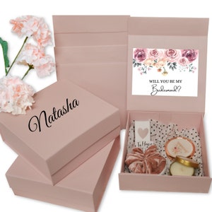 May include: Pink gift boxes with the name "Natasha" in cursive. One box is open, revealing a card that says "Will You Be My Bridesmaid?" and contains a scrunchie, candle, and other gifts. Artificial pink flowers are in the background.