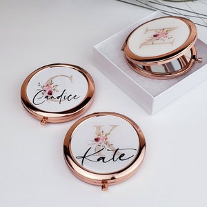 May include: Two rose gold compact mirrors with floral designs and personalized names. The mirror on the left says "Candice" and the mirror on the right says "Kate".