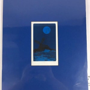 Puede incluir: Una impresión azul y negra de una luna sobre una pequeña isla con un árbol. La impresión está enmarcada en blanco y montada en un passe-partout azul.