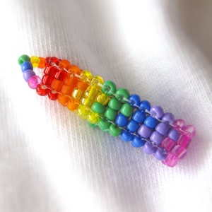 Peut inclure: Un porte-clés artisanal en perles aux couleurs de l'arc-en-ciel. Il est composé de petites perles colorées disposées en dégradé, passant du rouge à l'orange, au jaune, au vert, au bleu, au violet et au rose. Sur fond blanc.