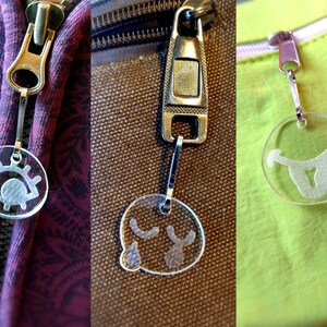 Puede incluir: Primer plano de tres tiradores de cremallera con colgantes circulares transparentes. Cada colgante presenta un gráfico blanco diferente: un ojo, una cara llorando y una cara sonriendo. Las cremalleras están unidas a telas en tonos morado, marrón y verde.