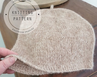 May include: A beige knitted hat with a small brim and a top loop. The image features text that reads JINXED STITCHES KNITTING PATTERN KNITWEAR DESIGNS and Intermediate. The hat is constructed with a stockinette stitch pattern and a garter stitch brim.