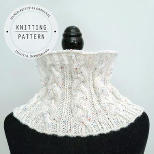 May include: A white knitted cowl with cable knit detailing and flecks of colour. The cowl is displayed on a black mannequin head. The image includes a circular graphic with the text "Jinxed Stitches Original Knitting Pattern Digital Download."