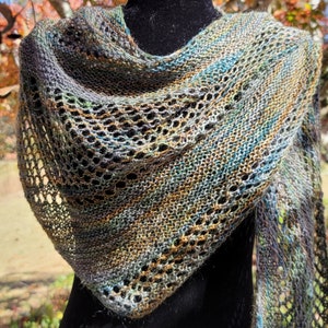 Puede incluir: Un chal tejido a mano en tonos grises, azules, verdes y dorados. El chal presenta un diseño calado y una forma triangular. El hilo parece ser una mezcla de colores, creando un aspecto texturizado y único. El chal está drapeado sobre una forma negra.