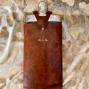 Può includere: Una fiaschetta rivestita in pelle marrone con un tappo argentato e un'iscrizione argentata che recita "H.L.G."