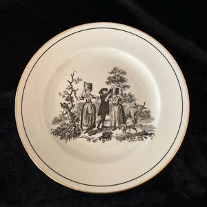 May include: A white ceramic plate with a black and gold rim. The plate features a black and white pastoral scene with figures, a dog, and a cow. The plate has a black line around the edge.