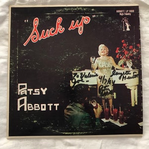May include: A black and white vinyl record cover with the title "Suck Up" in red lettering. The cover features a woman in a white dress holding a glass of liquid. The text "Patsy Abbott" is printed in white on a black background. The record is labeled "Abbott LP 1000 High Fidelity".