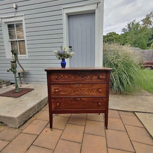 May include: A wooden dresser with three horizontal drawers, each featuring two round knobs and a central keyhole. The wood grain is visible on the drawer fronts, which are finished in a warm brown tone. A blue glass vase with flowers sits on top.