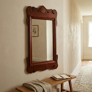 May include: A rectangular wall mirror with a dark brown wooden frame featuring a carved shell motif at the top center. The frame has decorative scrollwork details along the top and bottom edges, providing a structured and symmetrical appearance.