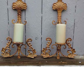 May include: A pair of gold-toned metal wall sconces featuring ornate scrollwork and a fan-shaped top. Each sconce holds a cylindrical off-white pillar candle. The metal has a distressed finish with visible texture and intricate leaf-like details.
