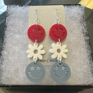 May include: A pair of dangle earrings with three charms. The top charm is a red smiley face with white stars for eyes. The middle charm is a white daisy. The bottom charm is a blue smiley face with white stars for eyes. The earrings are silver.