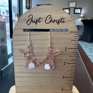 May include: Pink glitter bunny rabbit earrings with white fluffy tails on a wooden display stand with a ruler and the text "Just Christi" engraved on the top.