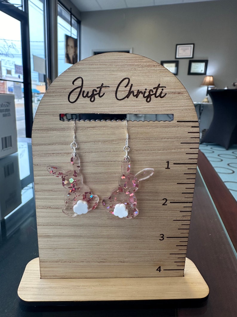 May include: Pink glitter bunny rabbit earrings with white fluffy tails on a wooden display stand with a ruler and the text "Just Christi" engraved on the top.