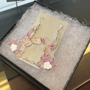 May include: A pair of silver earrings with a bunny design. The bunnies are made of clear resin with iridescent glitter and a white flower charm.