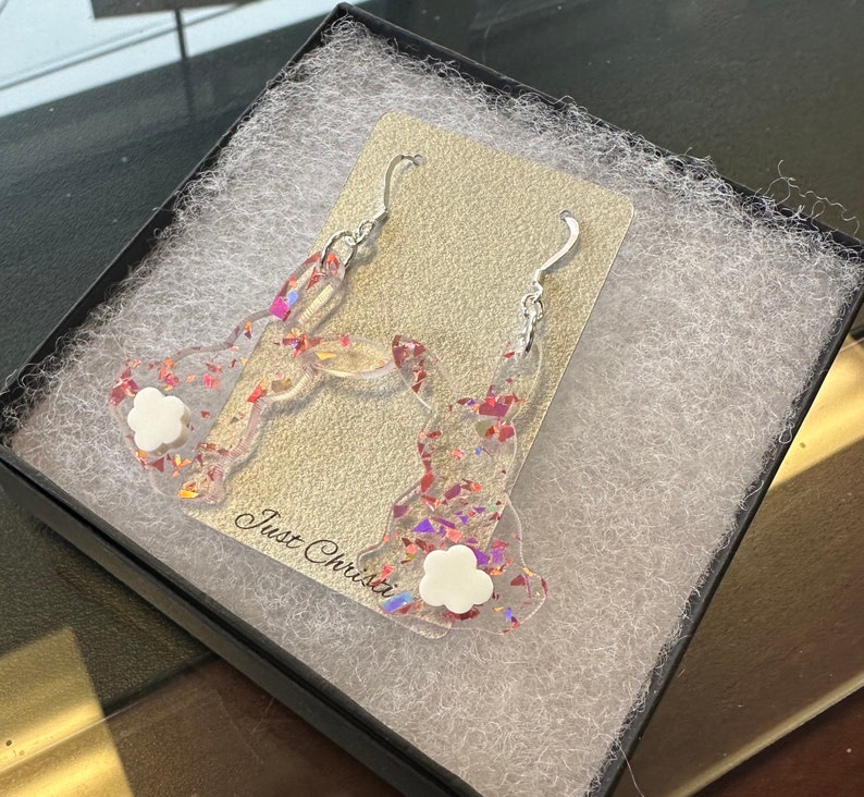 May include: A pair of silver earrings with a bunny design. The bunnies are made of clear resin with iridescent glitter and a white flower charm.
